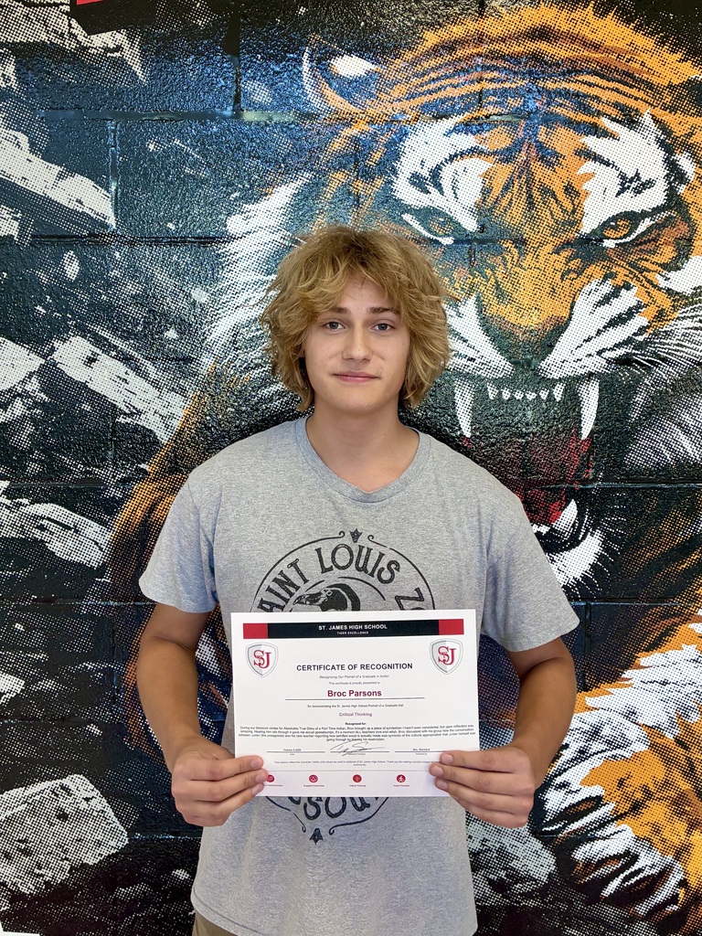 A young man holds a certificate in front of a tiger mural. He wears a gray T-shirt and has short blonde hair.