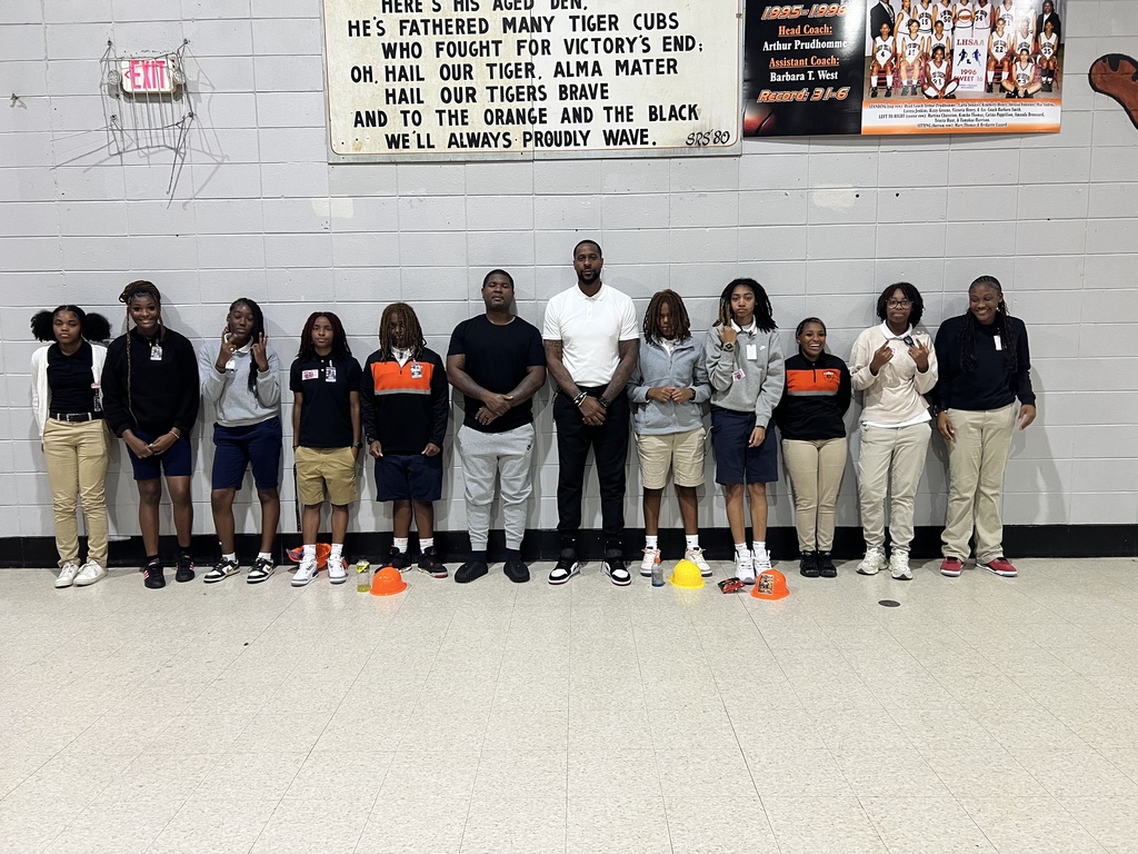 A group of Opelousas High School students stands with two adult guests in the school gym, dressed in school uniforms and OHS colors. Orange and yellow activity cones are on the floor in front of the group, with OHS history and memorabilia displayed on the wall behind them.