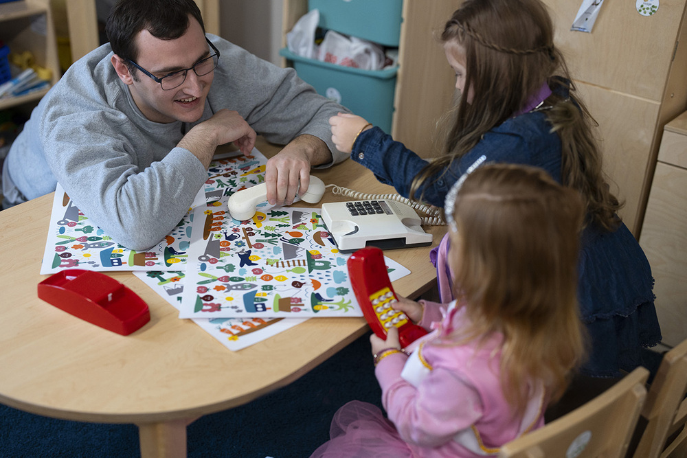 A preschool teacher sits at a table with two young children