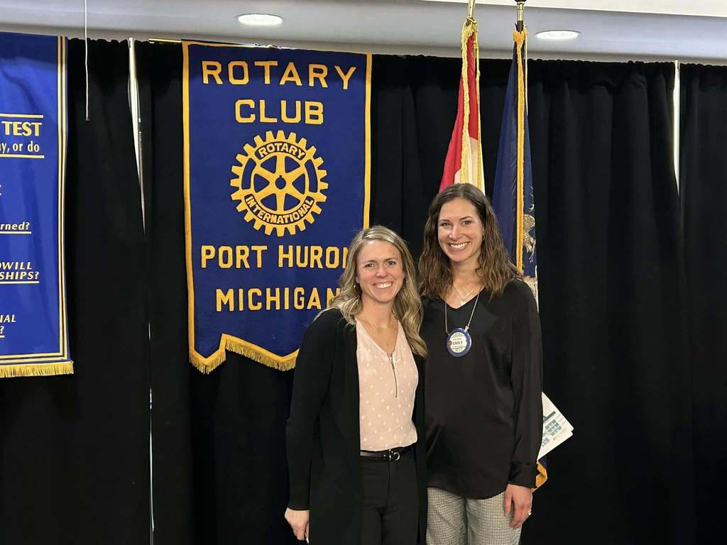 Two women recognized at Rotay Club Port Huron Michican. They stand in front of the American Flag and Rotay Club Flag.
