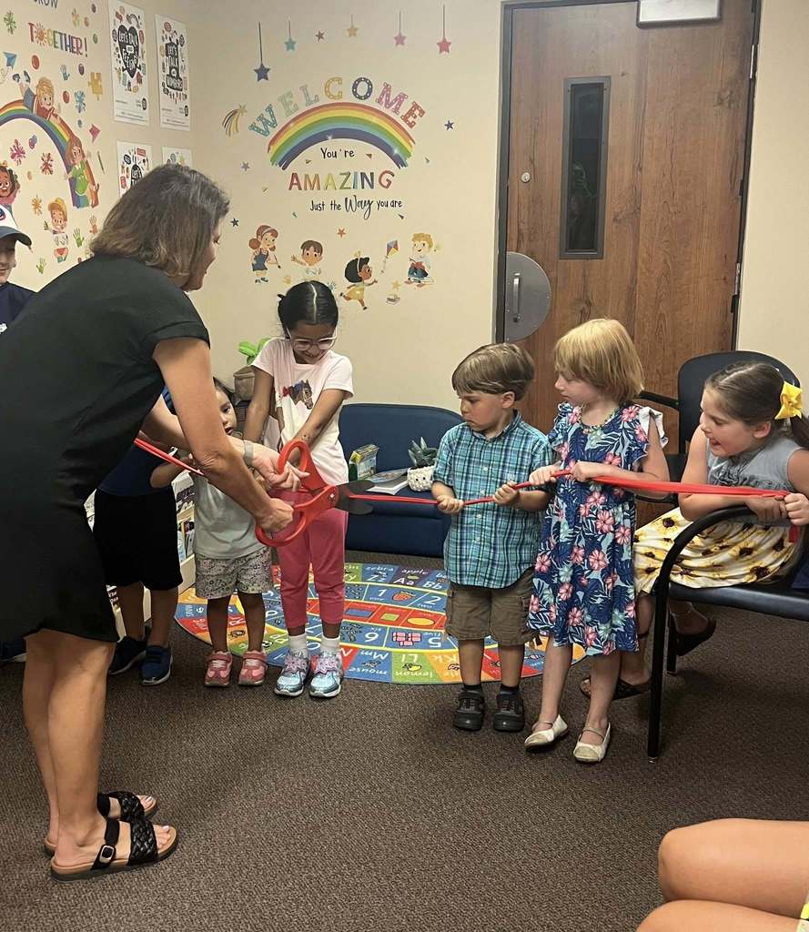 A group of children hold a red ribbon while an adult cuts it with a large pair of scissors.