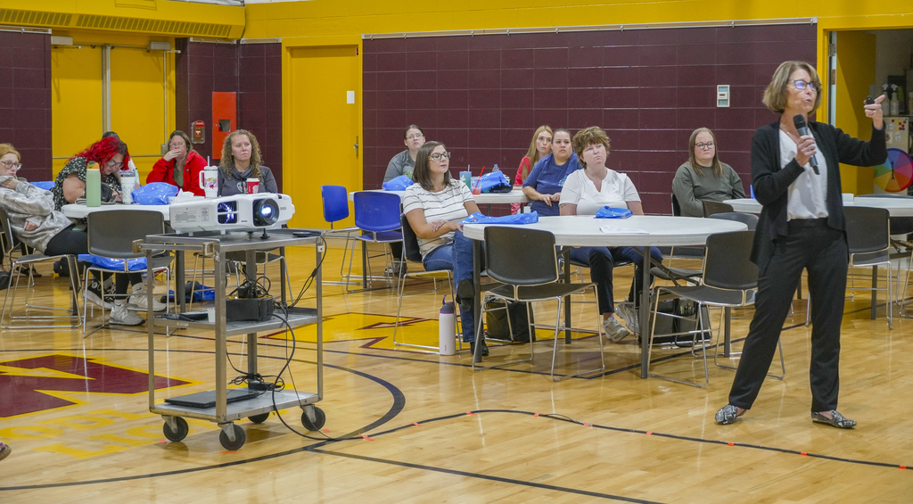 A presenter in a room full of people points to an electronic screen