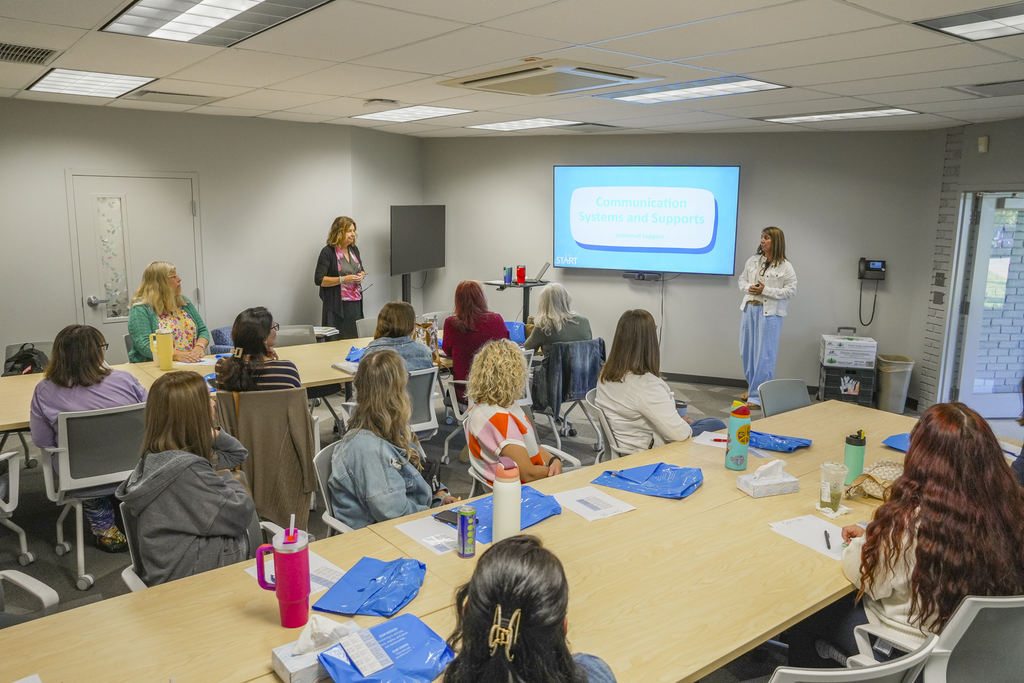 Two presenters stand in front of a screen presenting to a room of people