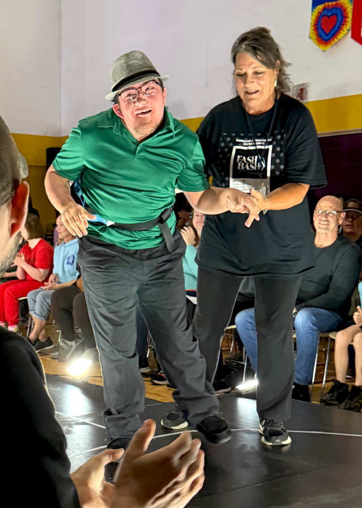 A smiling Woodland student wearing a green shirt, black pants, and a fedora is assisted down the runway by a Woodland staff member during Fash Bash