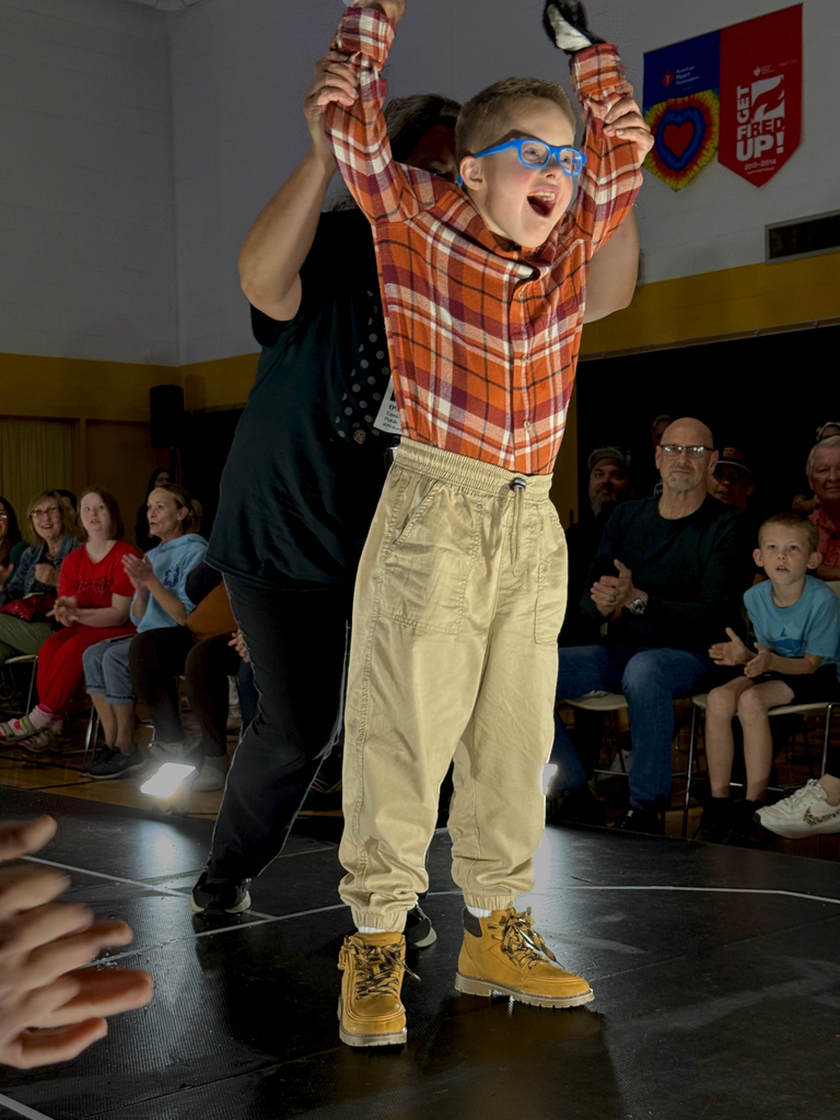 A young Woodland student modeling a plaid shirt, khakis, and work boots smiles as a Woodland staff member holds his arms in the air