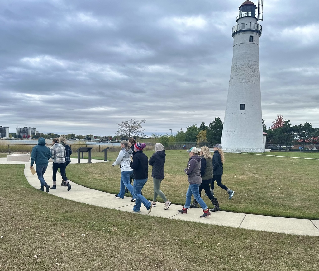 A small group walks along a paved path toward the Fort Gratiot lighthouse, bundled in jackets and hats on a cool fall day.