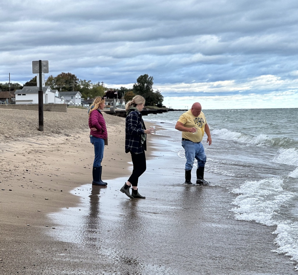 Three people in boots stand near the edge of the water, holding clear containers as they scoop samples from Lake Huron.
