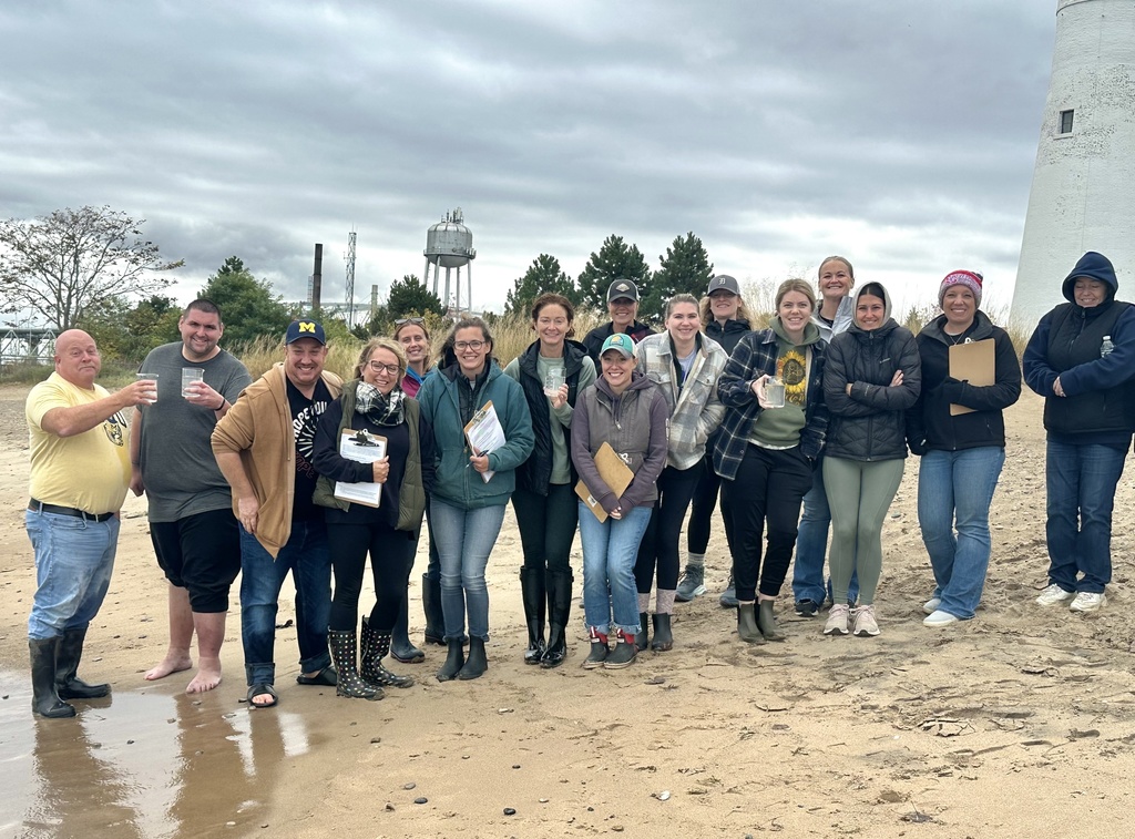 A group of Second Shift Academy participants stand smiling on Lighthouse Beach in Port Huron with the historic  lighthouse and the Blue Water Bridges visible in the distance