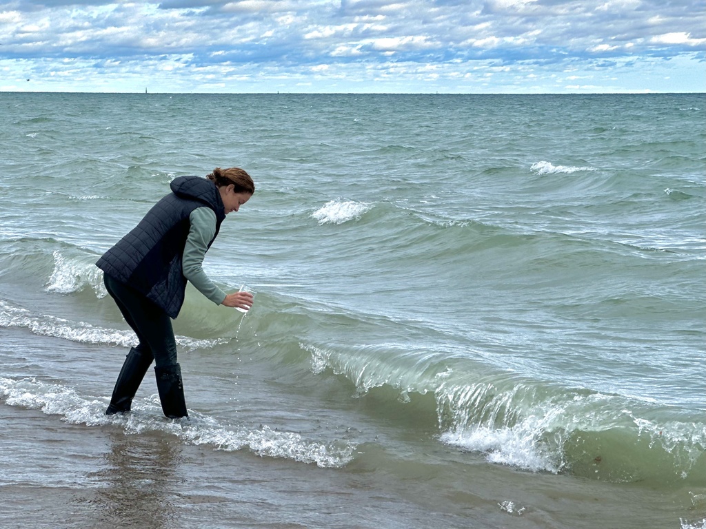 A participant leans toward the lake, collecting water in a clear cup as small waves crash on the shore.