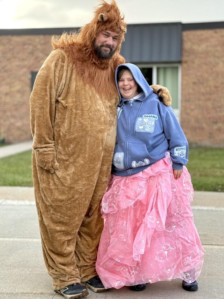 A person in a lion costume standing next to a person wearing a blue hoodie and pink princess skirt, both smiling.