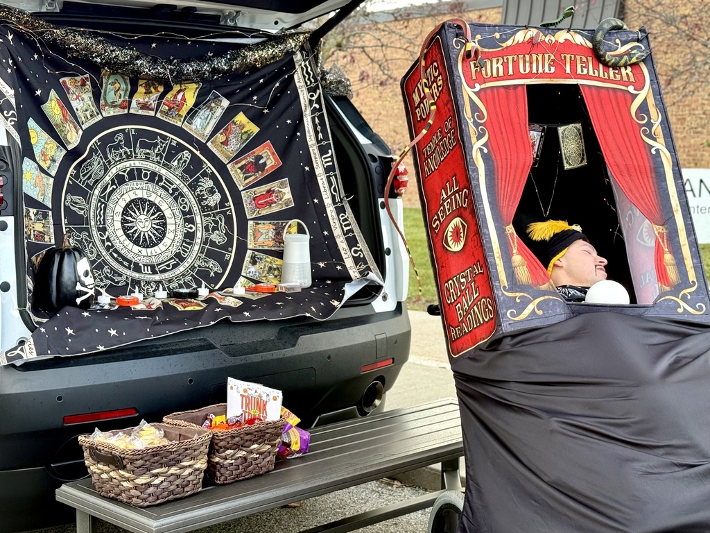 A car trunk decorated with a Zodiac and tarot card theme next to a person dressed as a fortune teller sitting inside a prop booth
