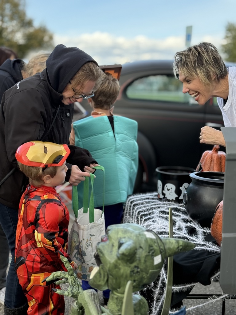 A woman laughing with a child dressed in an Iron Man costume as they collect candy at a spooky, web-decorated table.