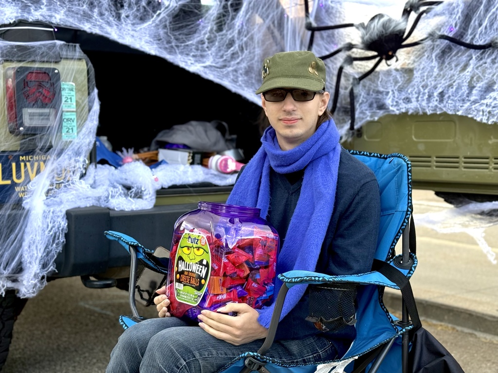 A person sitting in front of a truck decorated with fake spider webs and a large spider, holding a container of cheese ball snacks.