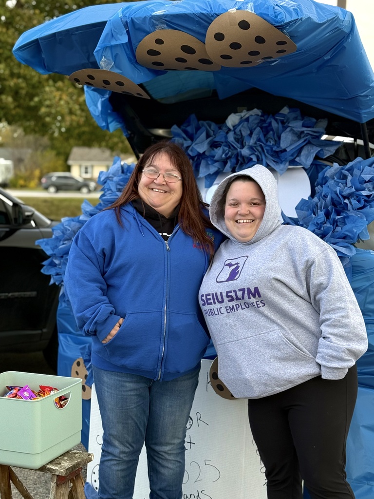 Two people in blue hoodies smiling in front of a trunk decorated with a Cookie Monster theme and large paper chocolate chip cookies.
