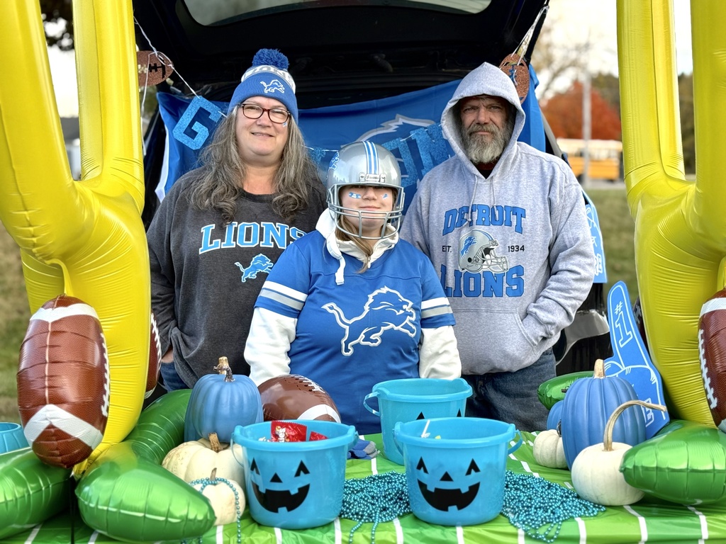 Three people dressed in Detroit Lions gear pose in front of a football-themed trunk with inflatable goalposts, pumpkins, and treat containers.