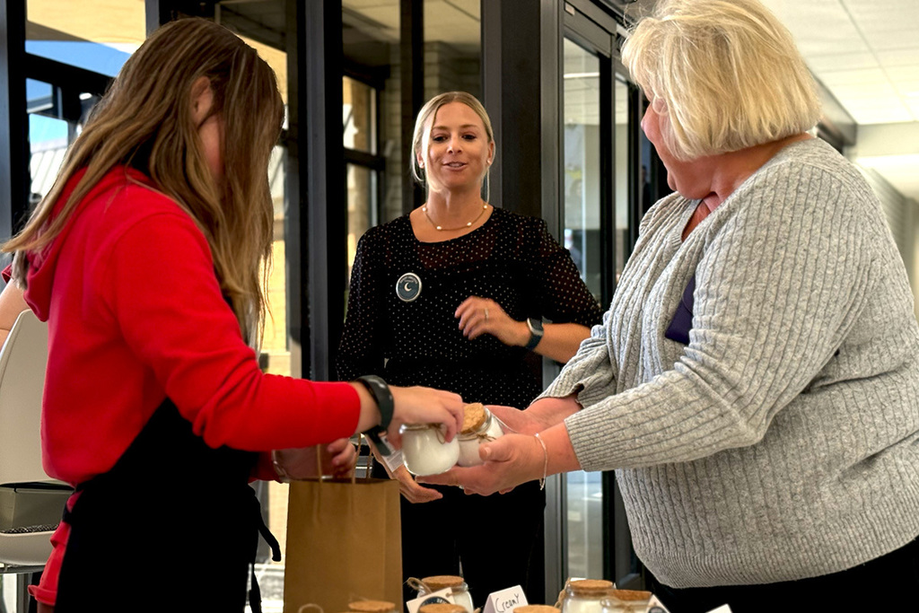 A student places a candle into a paper bag for a customer while teacher Next Step Karolynn Poff looks on and smiles.