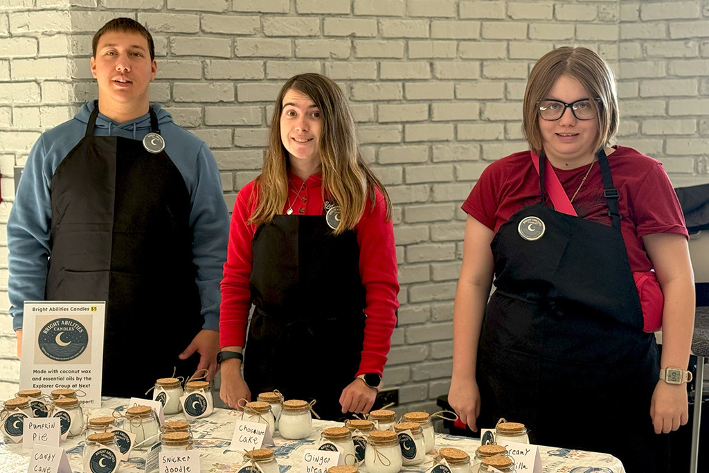 Three students wearing aprons stand behind a display of Bright Abilities Candles on a table, smiling for the camera. Each candle is labeled with a different scent and tied with twine.