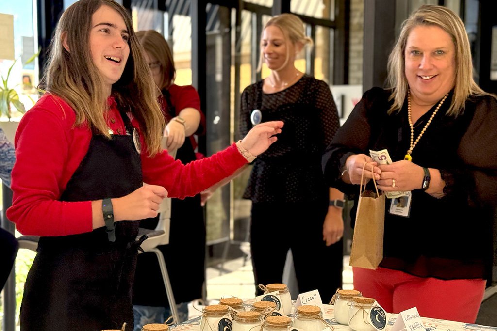 A student wearing a red shirt and black apron hands a bag to a smiling adult customer holding cash. Other students and staff are visible in the background.