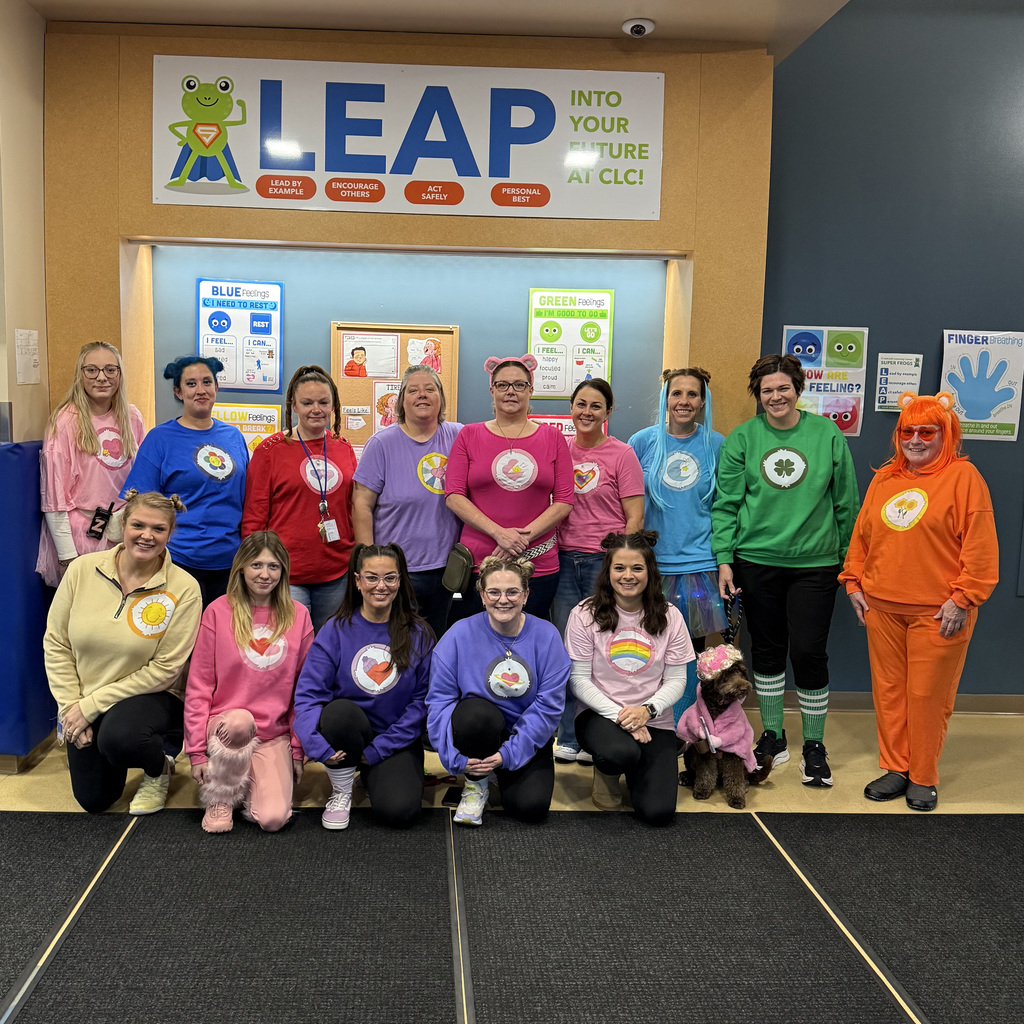Creekside Learning Center staff pose together in the school lobby wearing colorful Care Bear sweatshirts with Frannie the therapy dog sitting in front