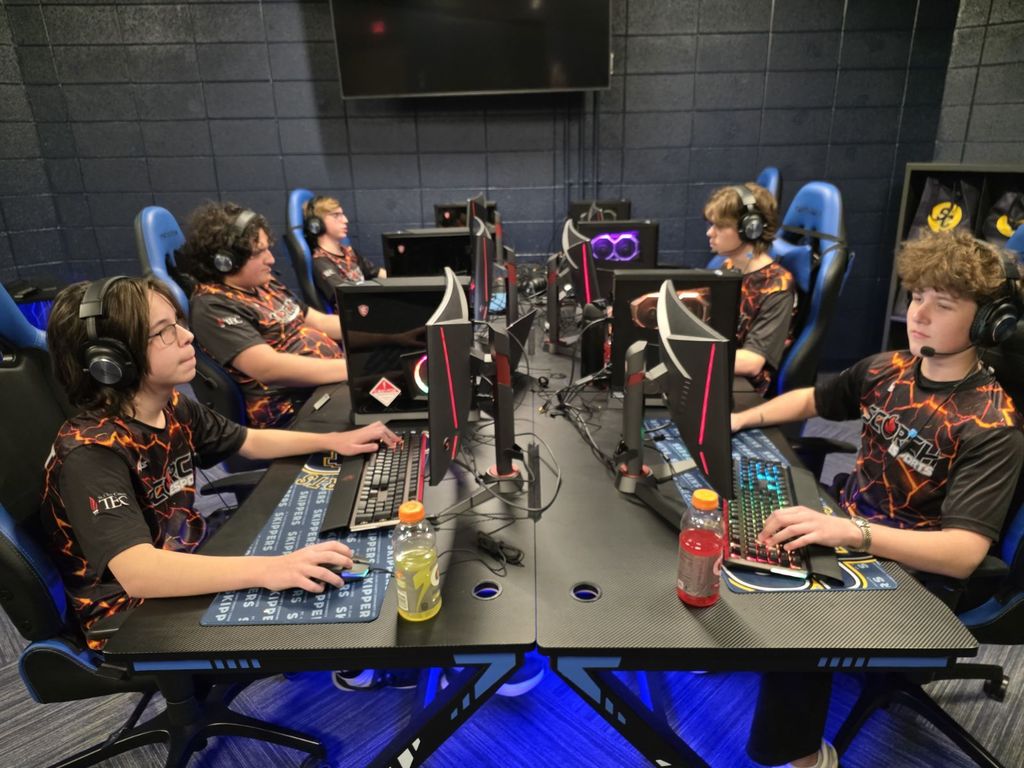 Five students wearing Scorch Esports jerseys sit at gaming desks in a dimly lit esports room. Each student wears a headset and plays on a desktop gaming computer with glowing lights. They face their monitors in two rows, focused on their match.