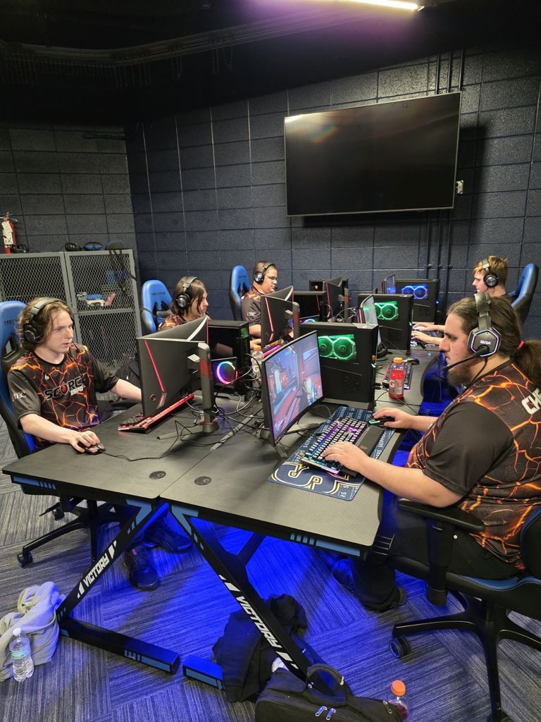 Five students in Scorch Esports jerseys sit side-by-side at high-performance gaming computers. They wear headsets and focus on their monitors while playing. Gatorade bottles and personal items sit on the desks as blue LED lighting glows beneath them.