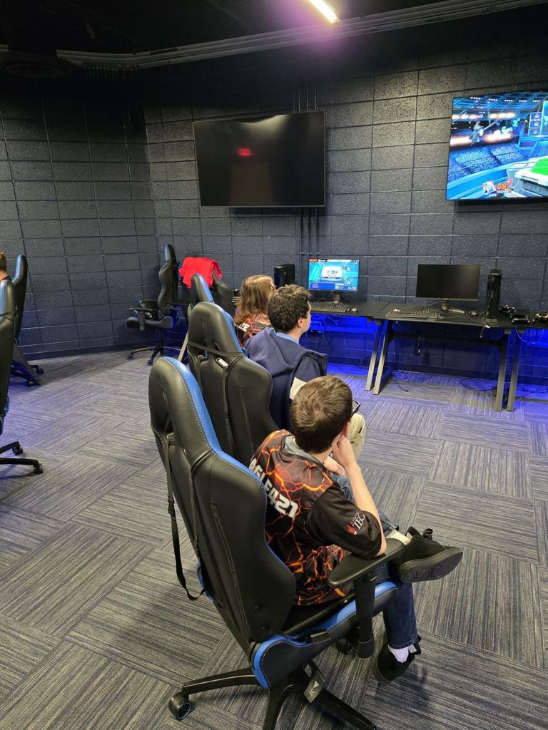 Three students, two wearing Scorch jerseys, sit in gaming chairs watching a Super Smash Bros. Ultimate match on a large wall-mounted screen. A smaller monitor and gaming setup sit in front of them in a blue-lit esports room.