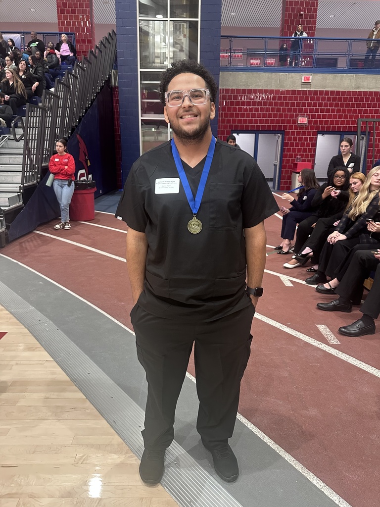 TEC student Braylon Rogan wearing black scrubs and glasses stands smiling with a gold medal around his neck. Other competitors and spectators are seated behind him in the gym.