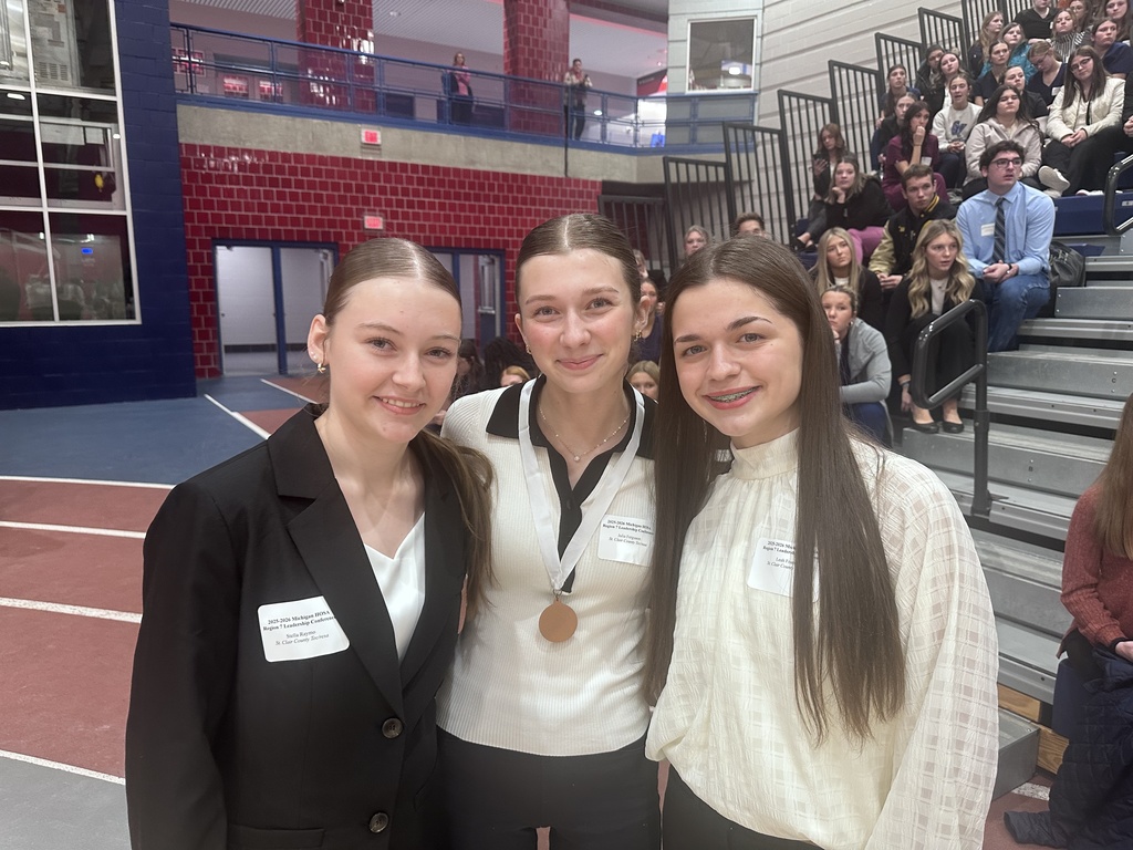 TEC students Stella Raymo, Julia Ferguson, and Leah Francek stand close together smiling. Bleachers of HOSA competitors are visible behind them.