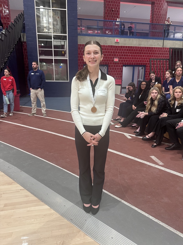 TEC student Julia Ferguson Wearing a white sweater and black pants stands smiling in the gym with a bronze colored medal around her neck. Students sit in the background. 