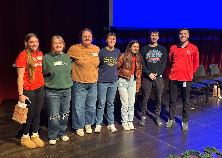 Six Port Huron High School alumni stand side by side on stage with BWCAN college advisor Garrett Goulding , smiling for a group photo before the Alumni Panel event.