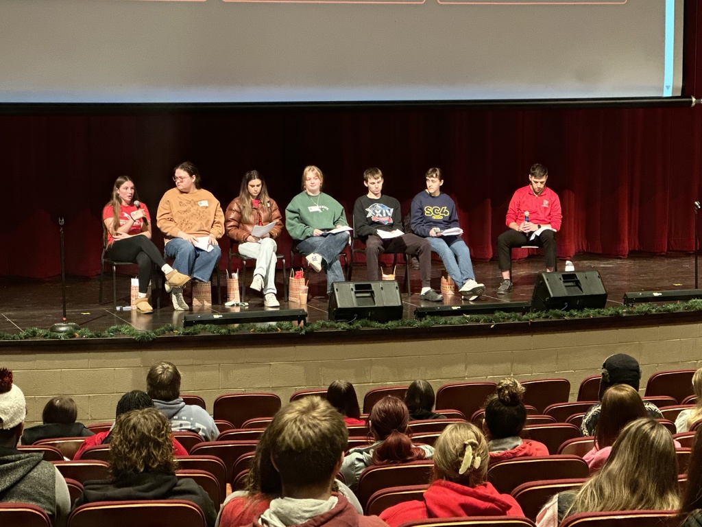 Six alumni and BWCAN college advisor Garrett Goulding sit in a row on stage holding notes and microphones while speaking to an audience of high school seniors.