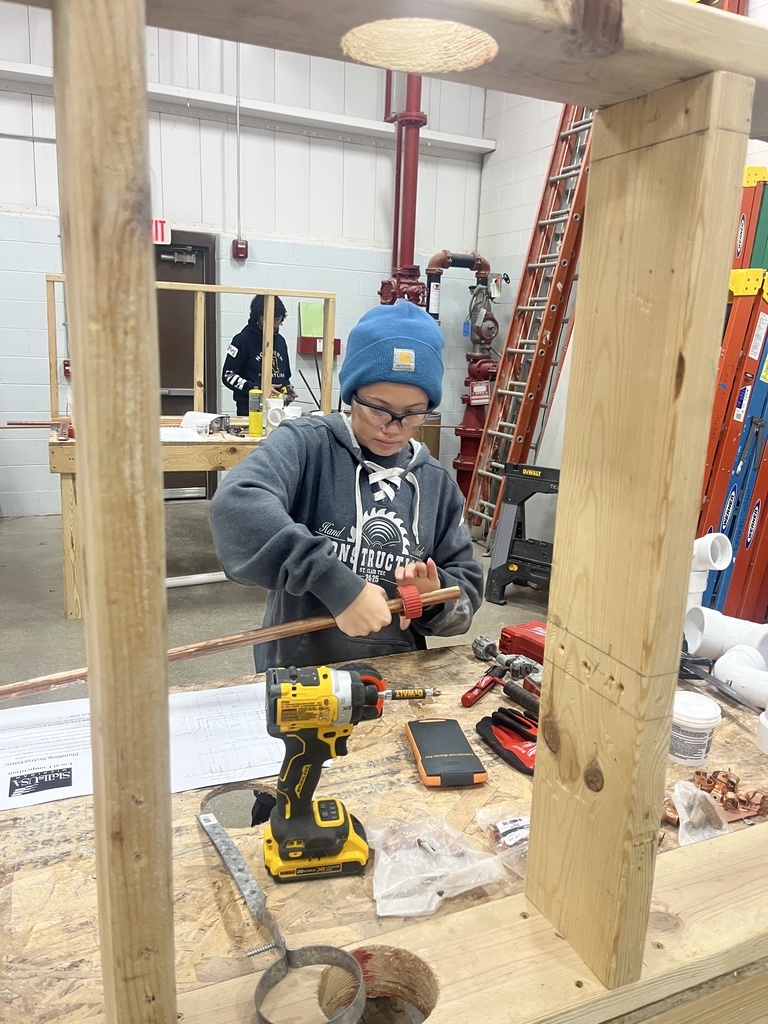 A student wearing safety glasses and a blue beanie uses a pipe-cutting tool on copper piping while working at a construction workstation 