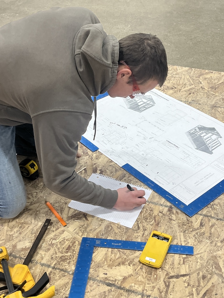 A student kneels on the floor reviewing a construction blueprints and writing notes, surrounded by measuring tools and equipment