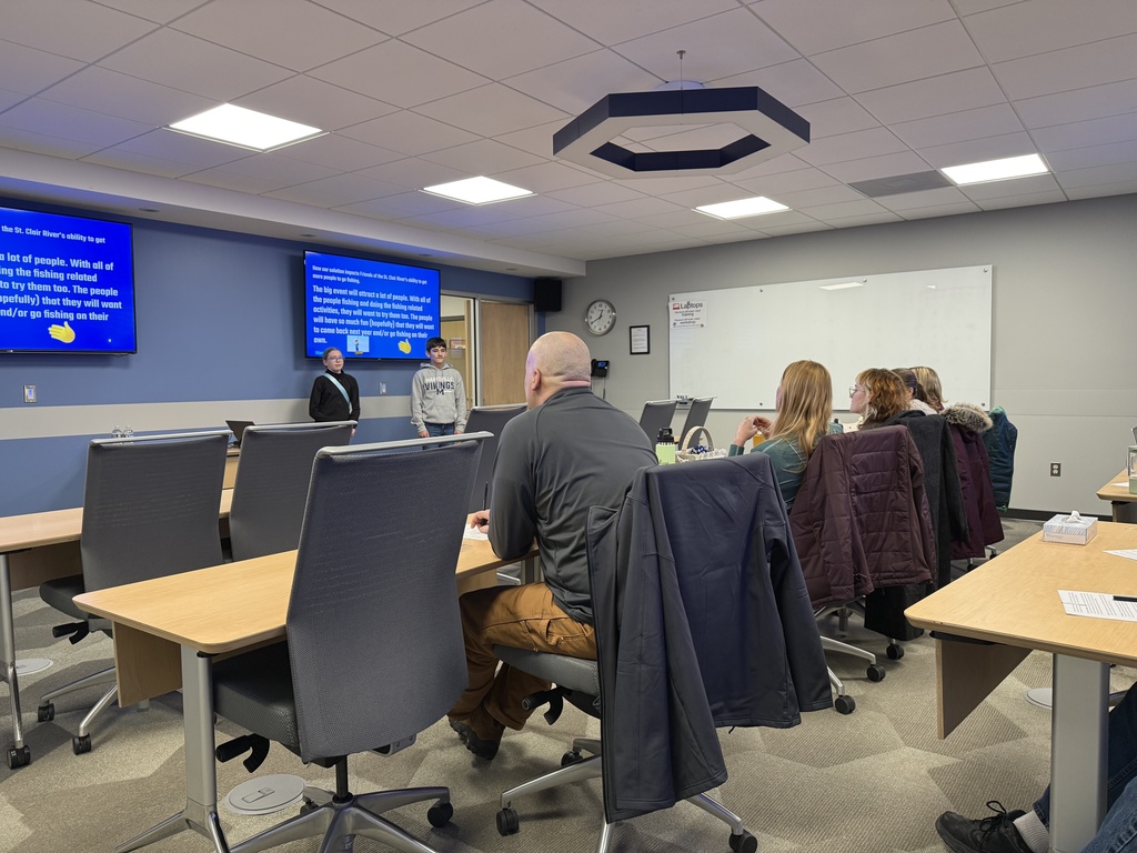 Two students stand at the front of a meeting room presenting to a group of adults