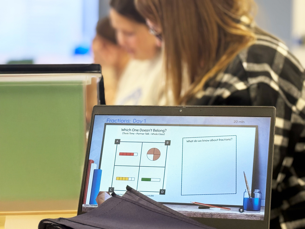 A laptop on a table displays a slide titled "Fractions: Day 1", showing fraction models and a prompt asking, Which one doesn't belong?"  