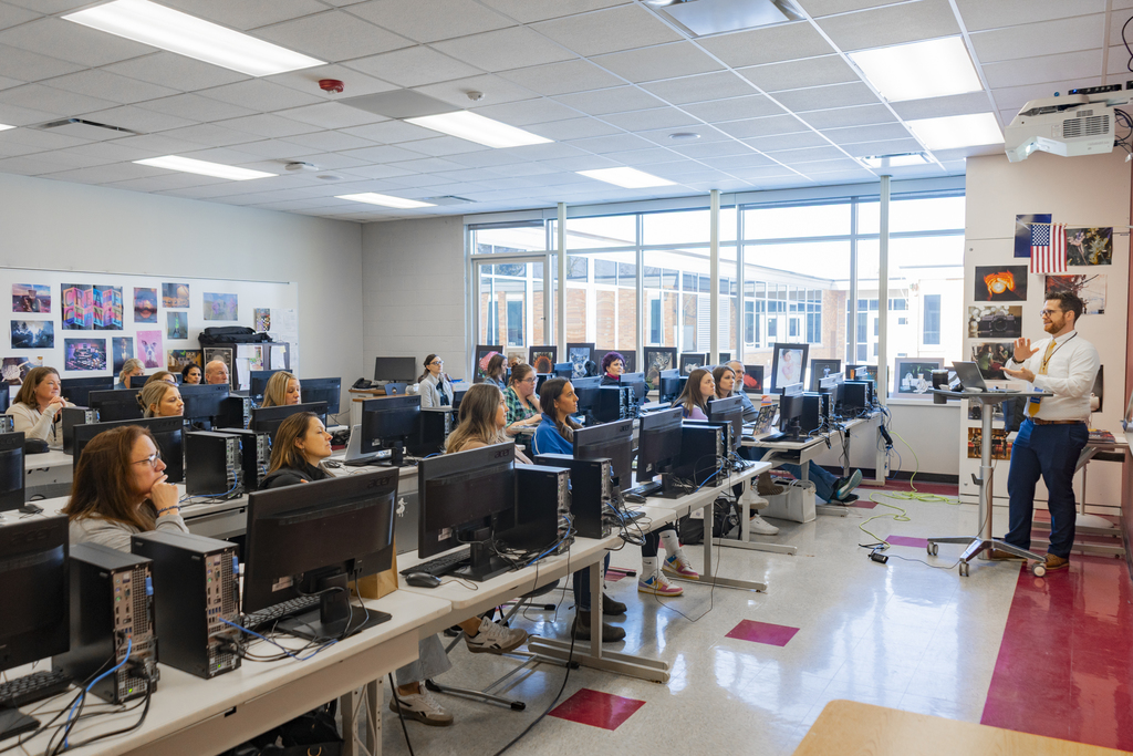 A presenter stands at a podium as he talks to a room full od people sitting at computers