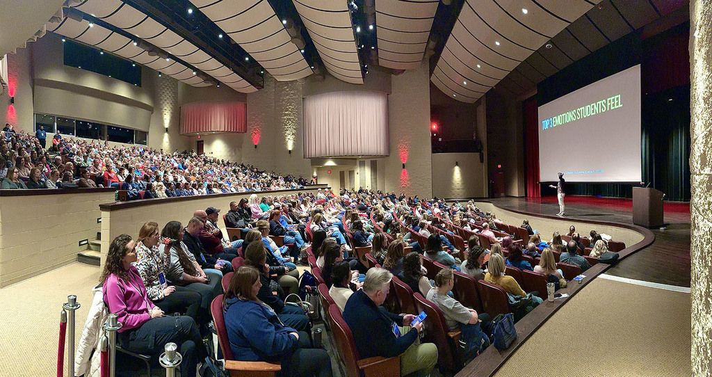 A speaker stands on the stage in a performing arts center in front of a full house