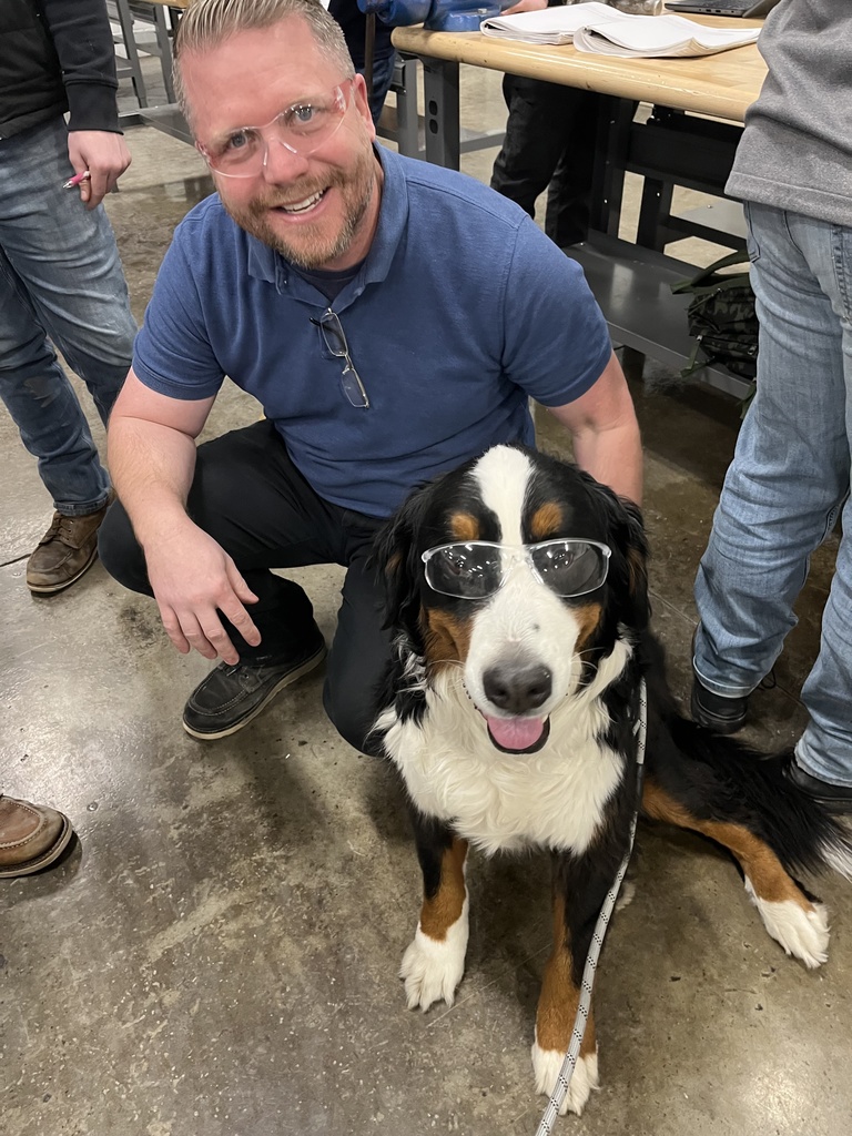 Automotive Technology instructor Jason Belfinger kneels on the shop floor next to Tank, a large Bernes Mountain dog. Both are wearing clear safety glasses and smiling at the camera. Work tables and other people are visible in the background.  