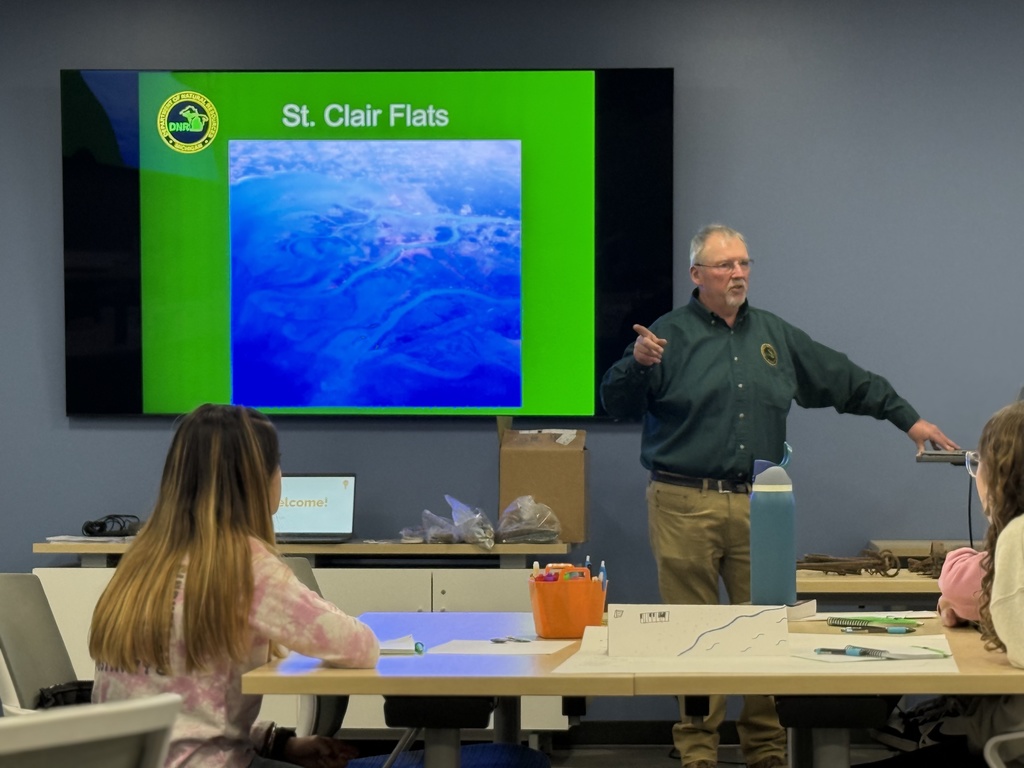A man stands in front of computer screen reading, "St Clair Flats," with students in the foreground.