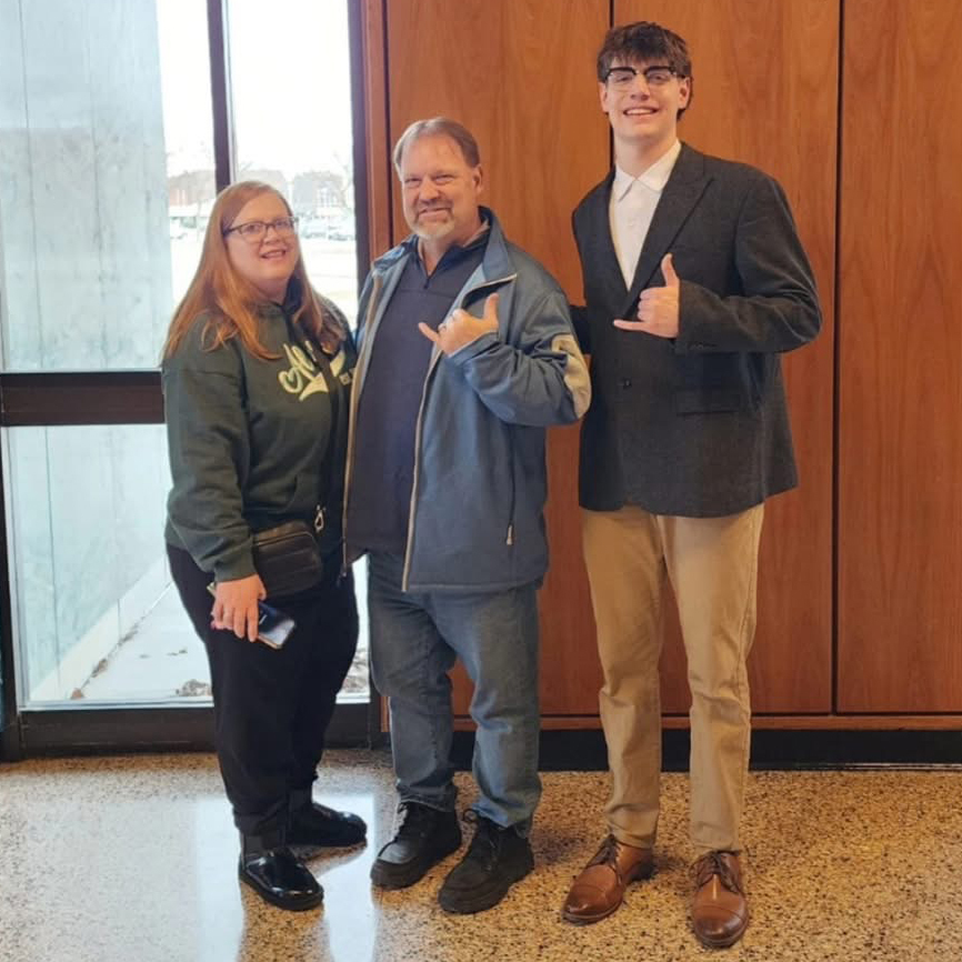 Three people stand together inside a building near a large window. The man in the center and the young man on the right both make a shaka hand gesture while smiling for the photo. 