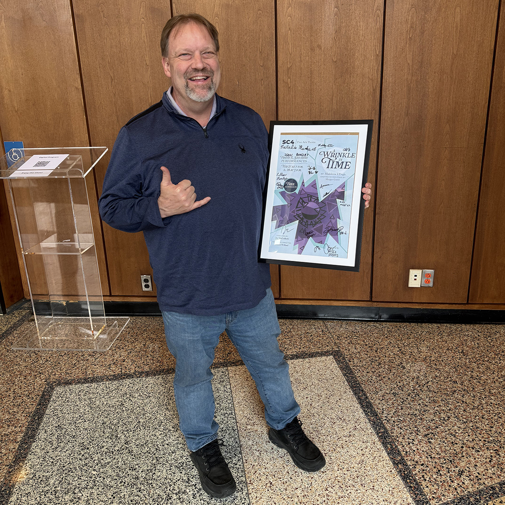 A smiling man stands indoors in front of a wood paneled wall holding a framed posted signed by the cast of "A Wrinkle in Time". He makes a shaka hand gesture wile posing with the poster. 