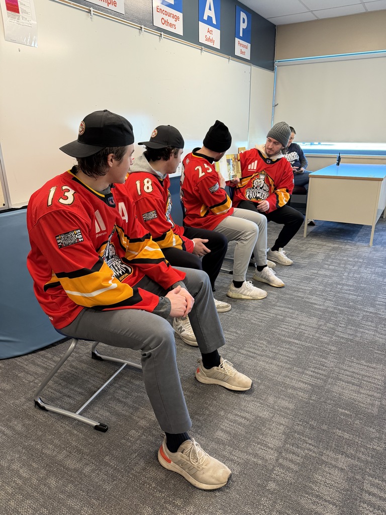 Four Port Huron Prowlers players wearing hats and hockey jerseys sit in a row at the front of a classroom. Two players hold open a picture book as they read to the class