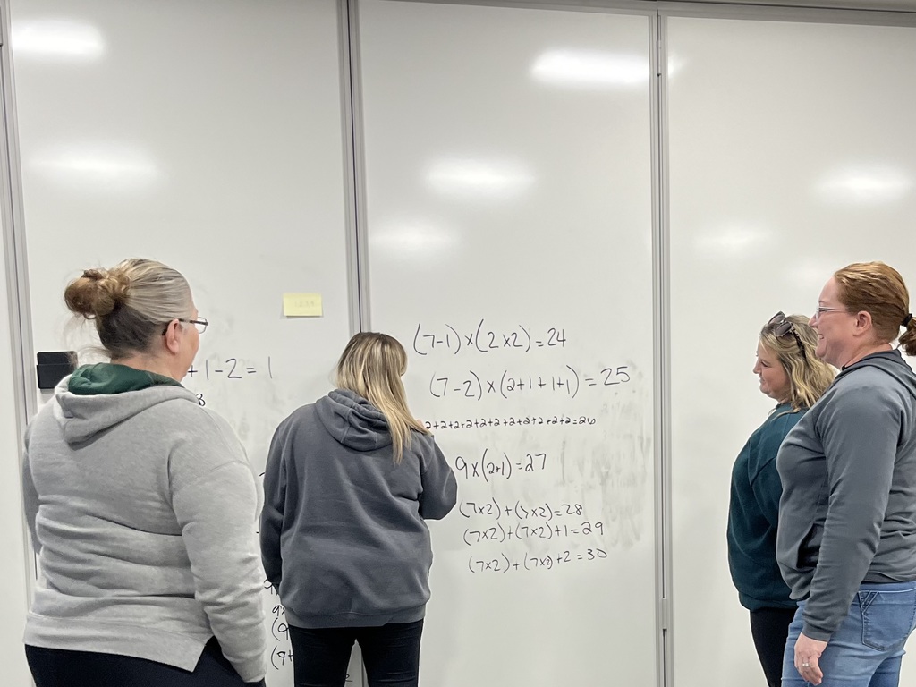 Four people with their backs to the camera stand at a whiteboard. One writes a math problem while the other three look on. 