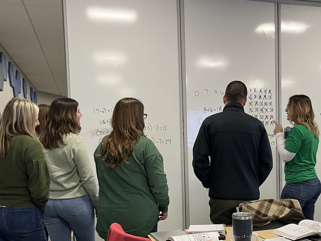 Six people with their back to the camera stand at a whiteboard. One person writes a math problem while the other five look on. 