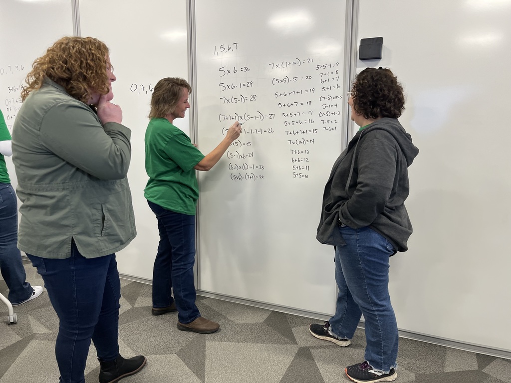 Three people stand at a white board. One writes a math problem while the other two look on.