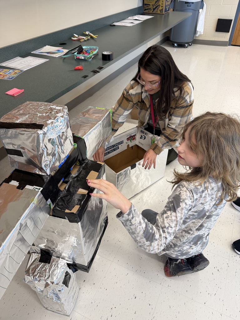 A student and teacher sit on the floor constructing a robot from recyclable materials.
