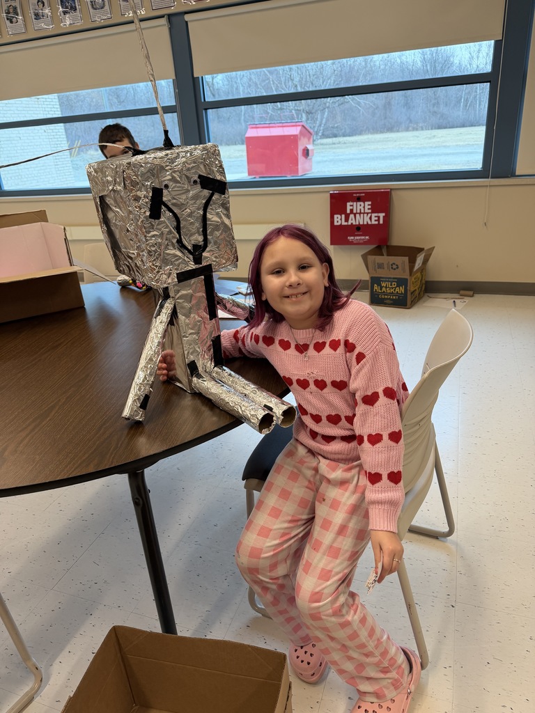 A student wearing a pink sweater and pants smiles at the camera with their arm wrapped around a robot constructed of boxes, paper tubes, and tinfoil