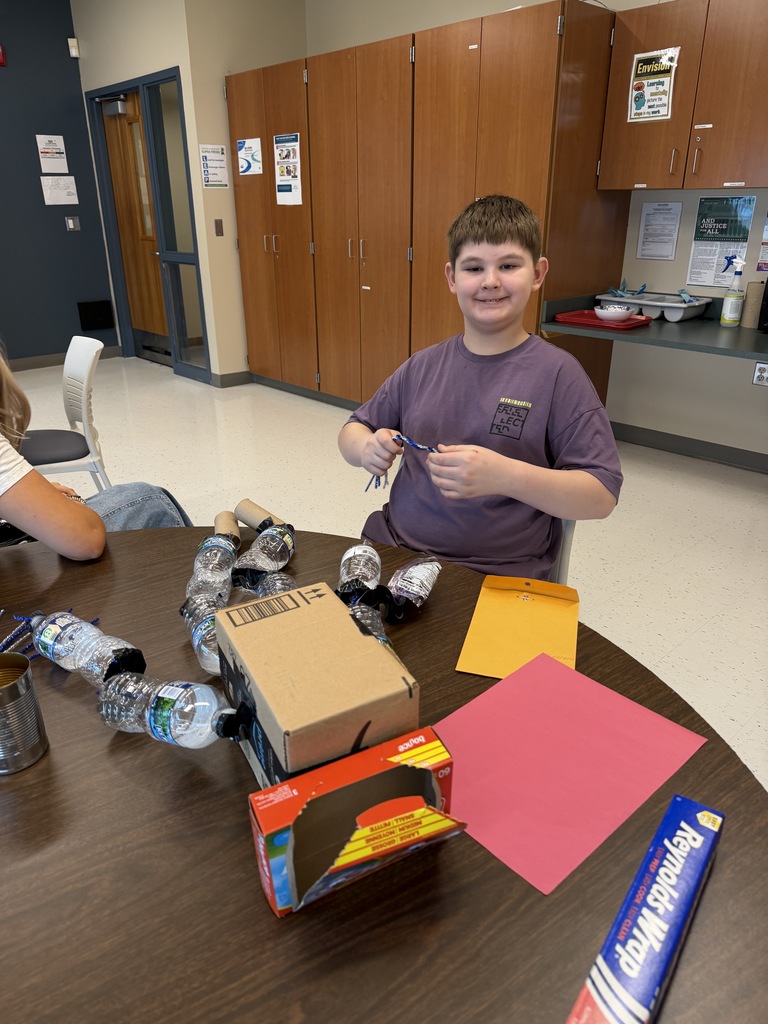 A student seated at a table smiles at the camera. They table holds materials such as cardboard boxes, plastic water bottles, paper tubes, colored paper, and tin foil. 