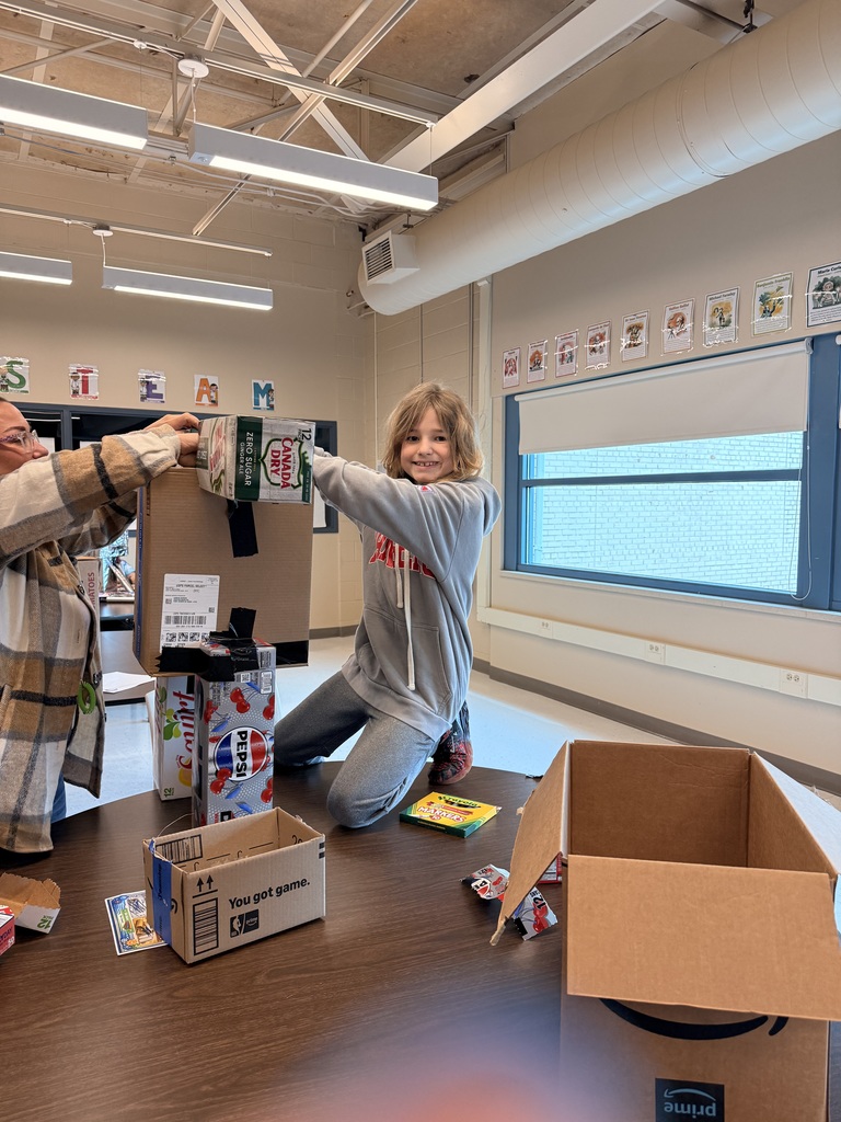 A student and teacher construct a robot using empty soda can boxes, markers, and cardboard boxes.