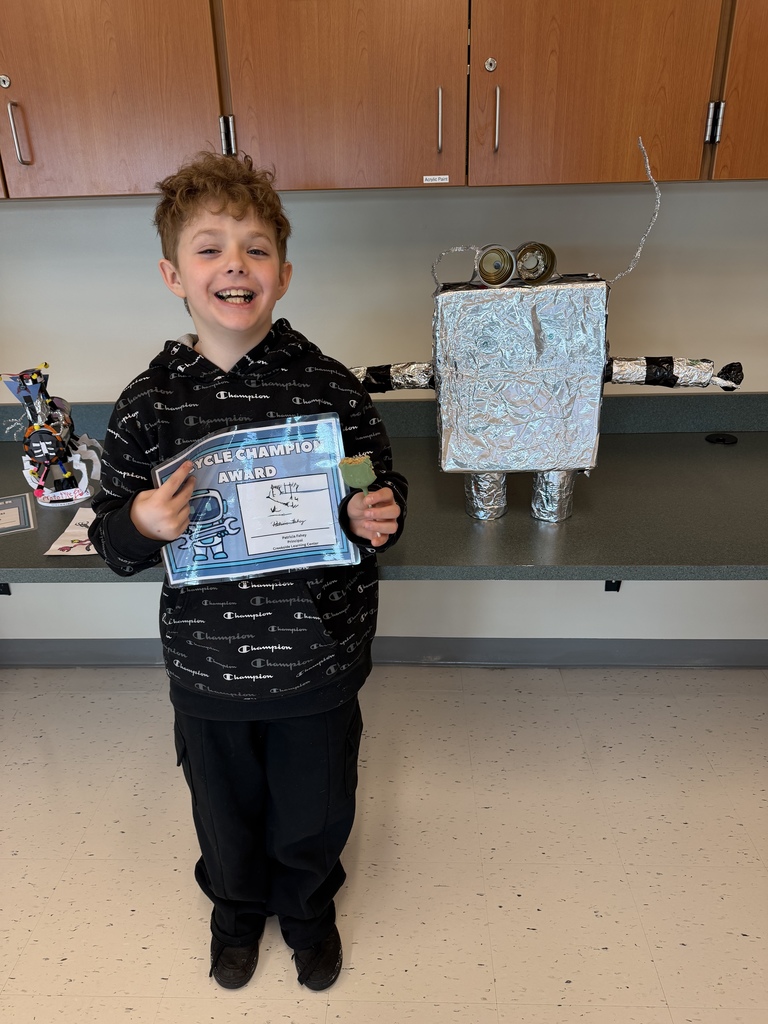 A student holding a certificate smiles at the camera as they stand next to their robot featuring two cans for eyes, and a tinfoil body